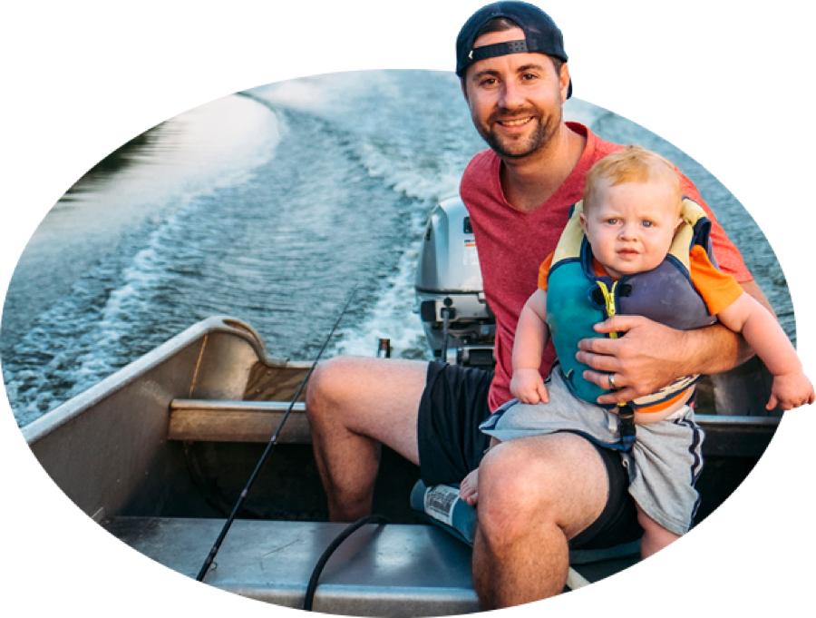 A bearded young father and his life-jacket-wearing toddler 电动机 across the water in a fishing boat, one of the many types covered by 国营农场 boat insurance.